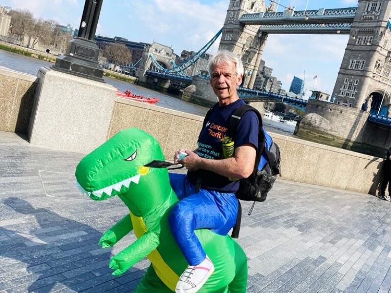 Geoff in a Guy's Cancer T shirt, bottom half hes got a green t-rex dinosaur costume in front of tower bridge.