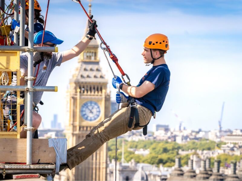 A Guy's Cancer Charity supporter at the top of the roof at St Thomas' hospital, about to abseil down. They're all wearing navy t-shirts that read 'Guy's Cancer Charity' on the front and wearing yellow helmets. Behind them is the iconic Big Ben.