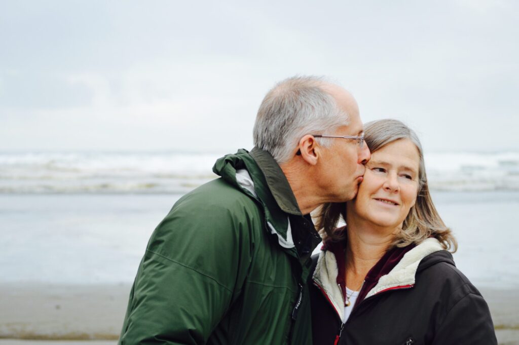 An elderly couple with the beach in the background, on the left the man is kissing his partner on her cheek and she is on the left looking away into the distance. Both are in waterproof coats.