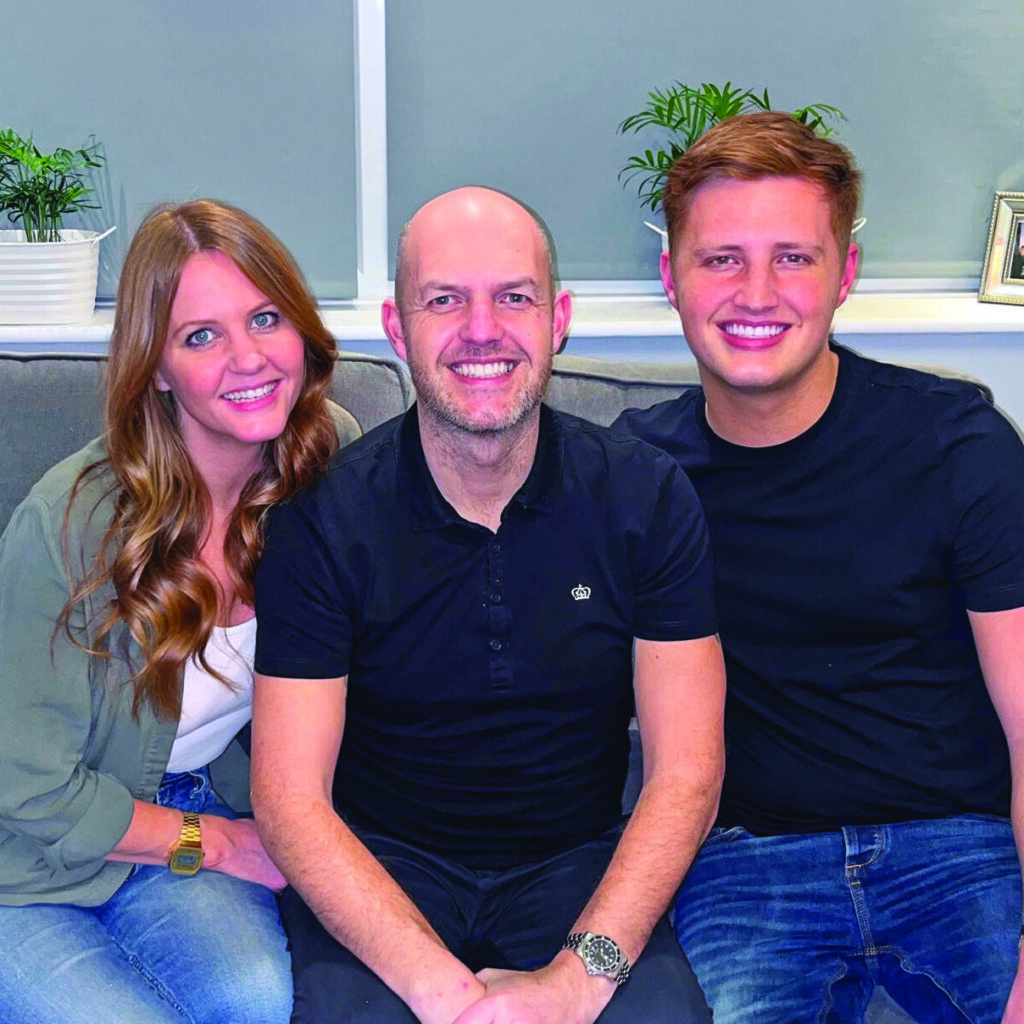 Alison, Gary and their son sat on a grey sofa. Behind them is a windowsill with two small green plants and a metal photo frame on the right.