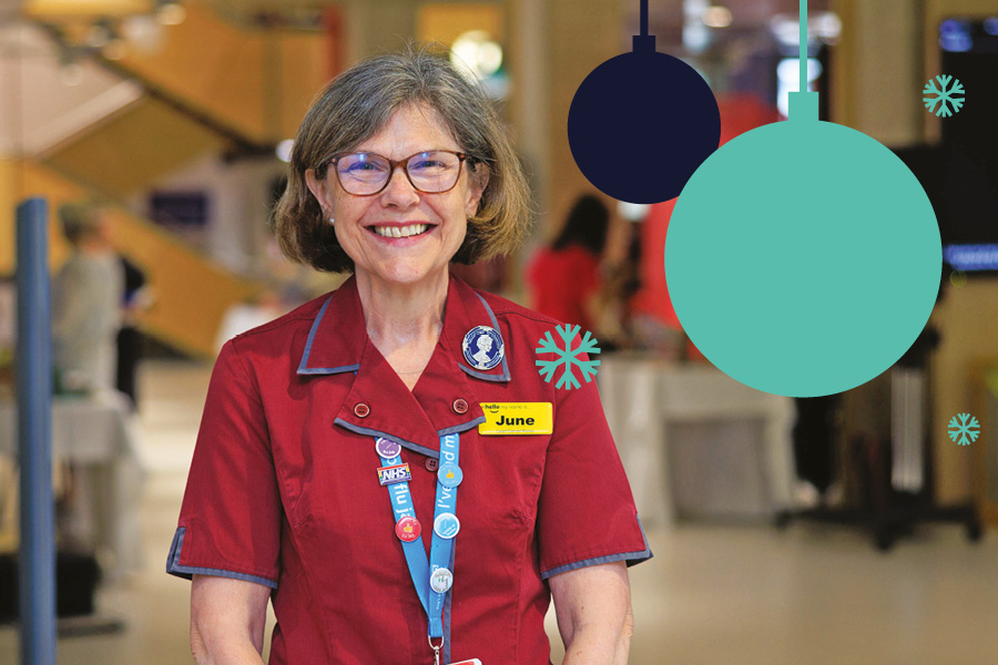 Nurse June, in glass and red uniform smiling in a hospital wearing a yellow name badge that reads 