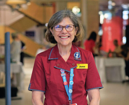 Nurse June, in glass and red uniform smiling in a hospital wearing a yellow name badge that reads 