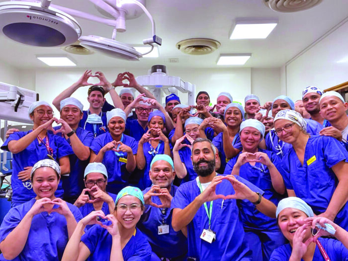 Group photo of 27 staff involved in high intensity theatre using their hands to do a heart shape symbol, all staff are wearing purple gowns and hair covers.