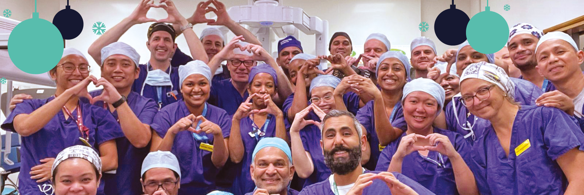 Group photo of 27 staff involved in high intensity theatre using their hands to do a heart shape symbol, all staff are wearing purple gowns and hair covers with blue and teal blue graphic baubles with teal snowflakes on the top.