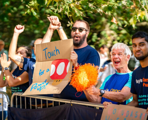 A group of supporters cheering on runners at an event, holding signs and pom-poms. One person is holding a cardboard sign that reads "Touch for a Power Up!" with a red mushroom illustration from Mario. Others are wearing shirts supporting Guy’s Cancer Charity and clapping. The scene captures a moment of joy and excitement as they cheer on the event