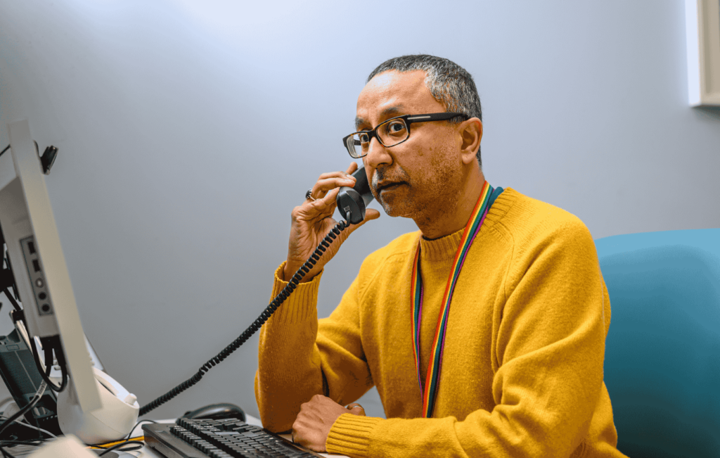 Nazim sat in a yellow jumper with a rainbow lanyard by his desk appearing to be taking a call on a spiral wired dark coloured phone. On his desk he