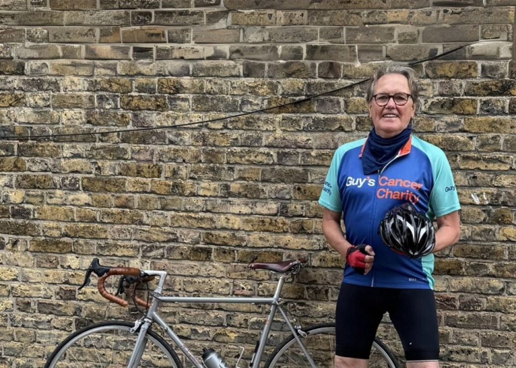 Dave pictured in front of a grey brick wall standing next to his bike which you can see the top of. Dave is wearing a blue Guy