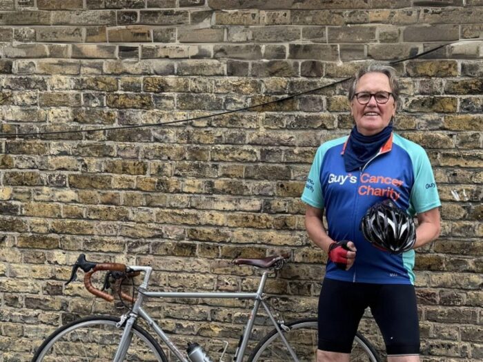 Dave pictured in front of a grey brick wall standing next to his bike which you can see the top of. Dave is wearing a blue Guy