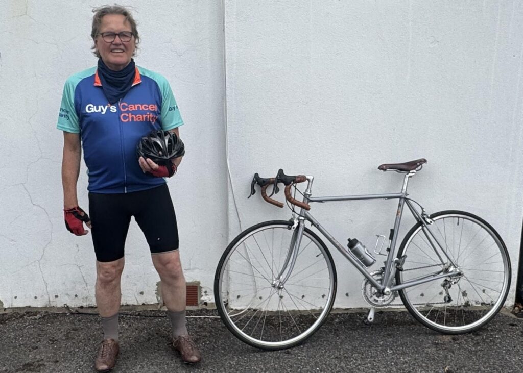 Dave pictured in front of a white wall standing next to his bike, Dave is wearing a blue Guy