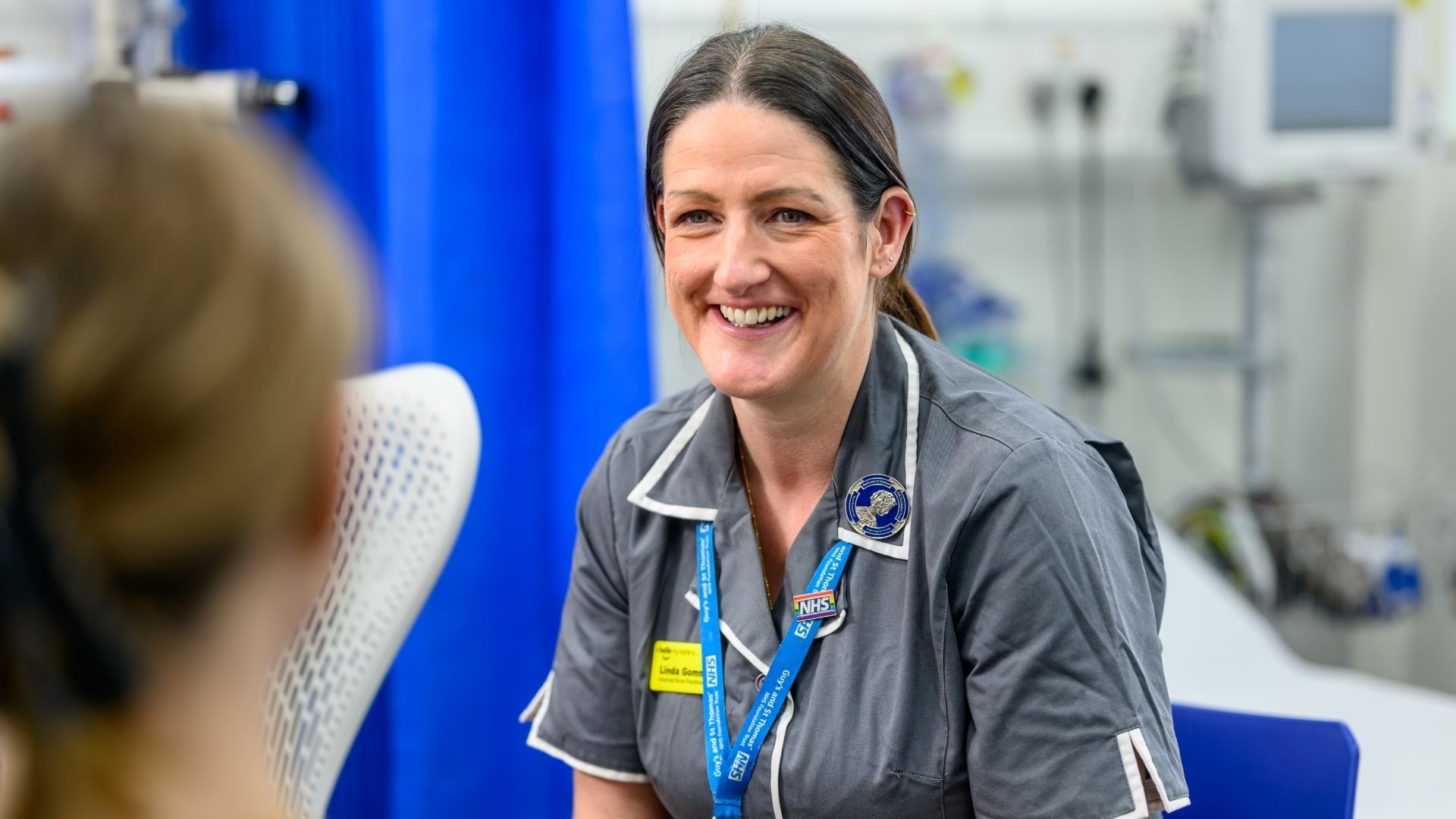 Guy's Cancer Nurse with brown hair tied back, wearing a grey short sleeve uniform appears to be smiling at a patient.
