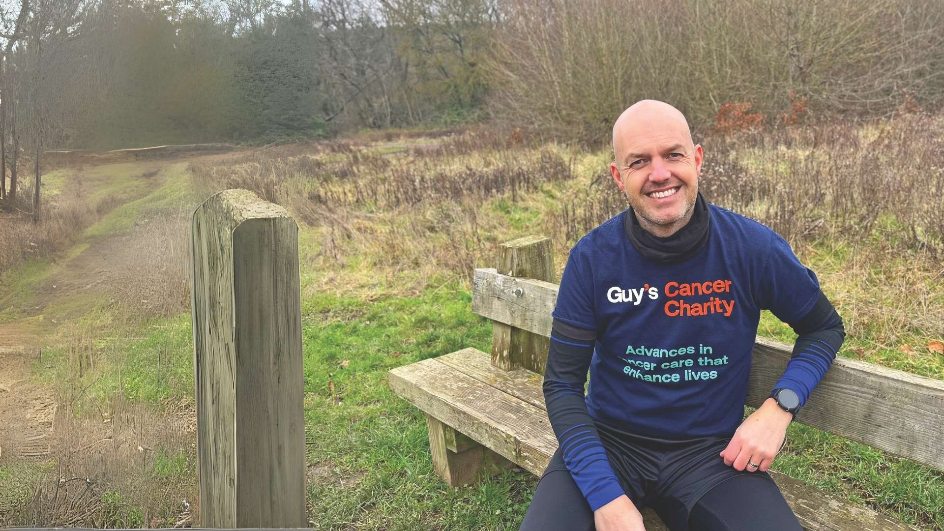 Gary sat on a wooden bench, wearing a navy t-shirt that reads 'Guy's Cancer Charity, advances in cancer care that enhance lives' with black and blue pattern long sleeve thermals underneath.