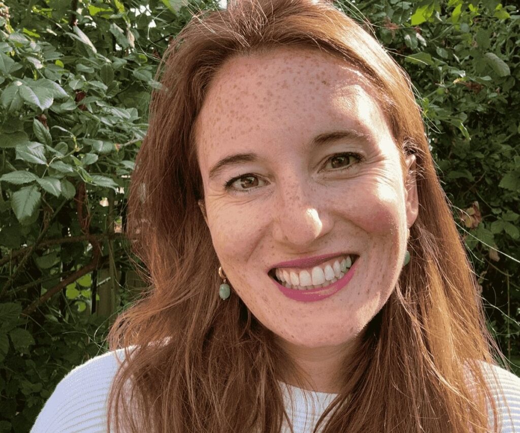 Portrait of Clare smiling, with long hair and leafy hedge in the background.