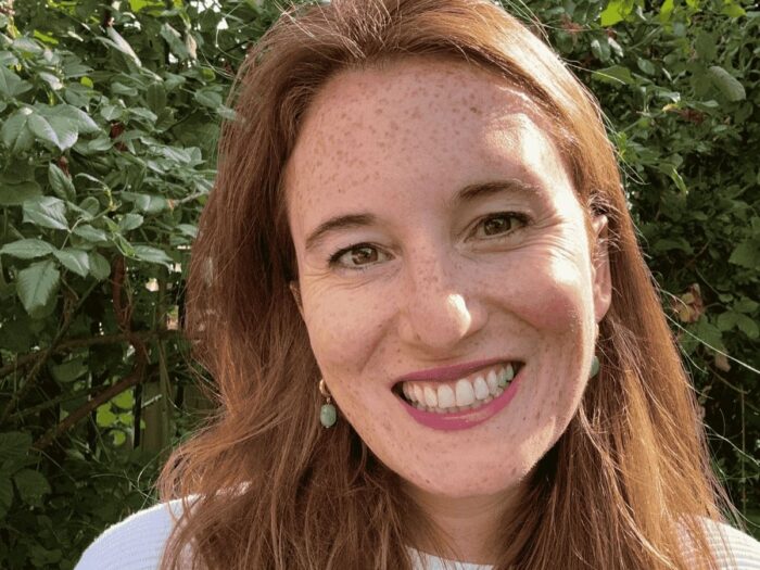 Portrait of Clare smiling, with long hair and leafy hedge in the background.