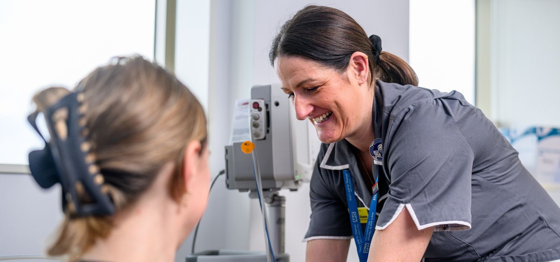 Guy's Cancer staff member taking care of a patient, she's in grey uniform with a blue lanyard, behind her is machinery and in front of her is the back of a patient.